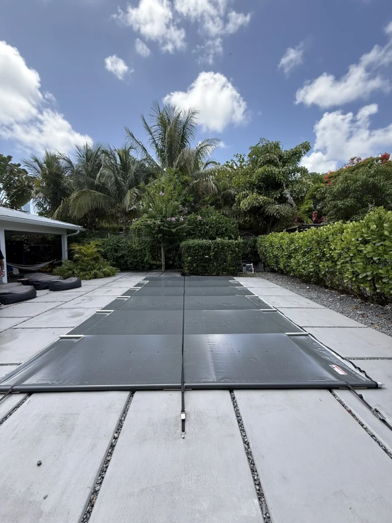 B-COVER manual safety pool cover installed over a symmetrical backyard pool in Miami, framed by palm trees and hedges under a blue sky