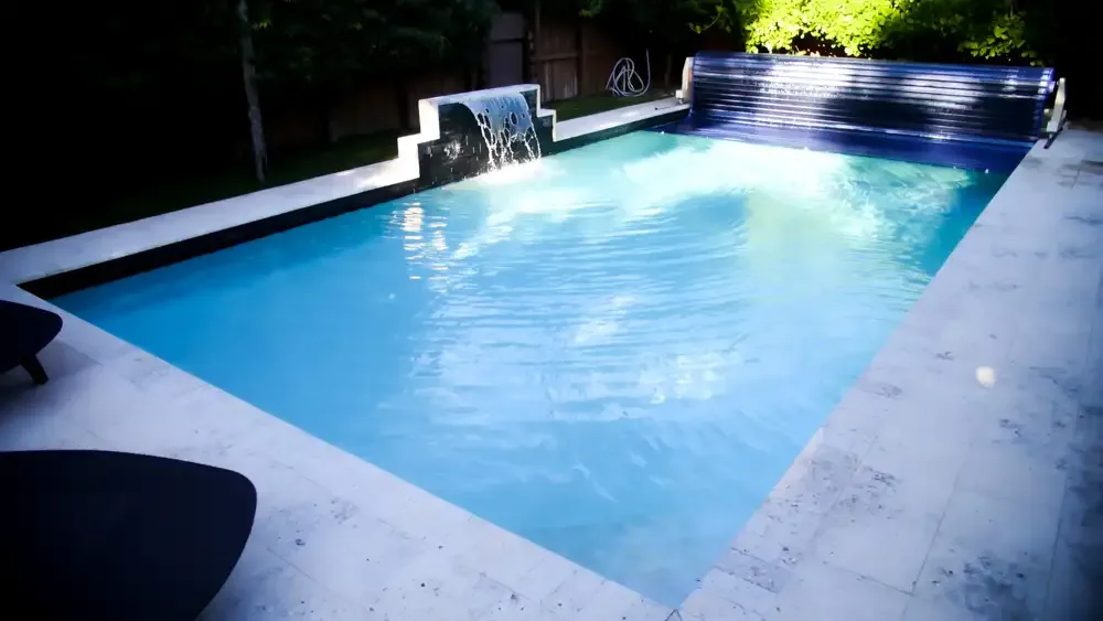 S-ROLL slatted automatic pool cover rolled open beside a lit pool with a water feature at night